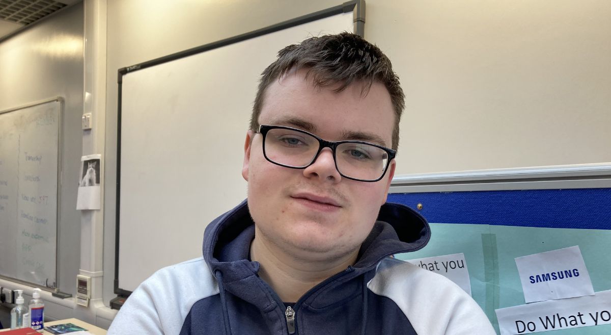 Ryan Johnston, young male with short dark hair, wearing glasses, a blue and white top and pictured in a classroom with notice board in background. 